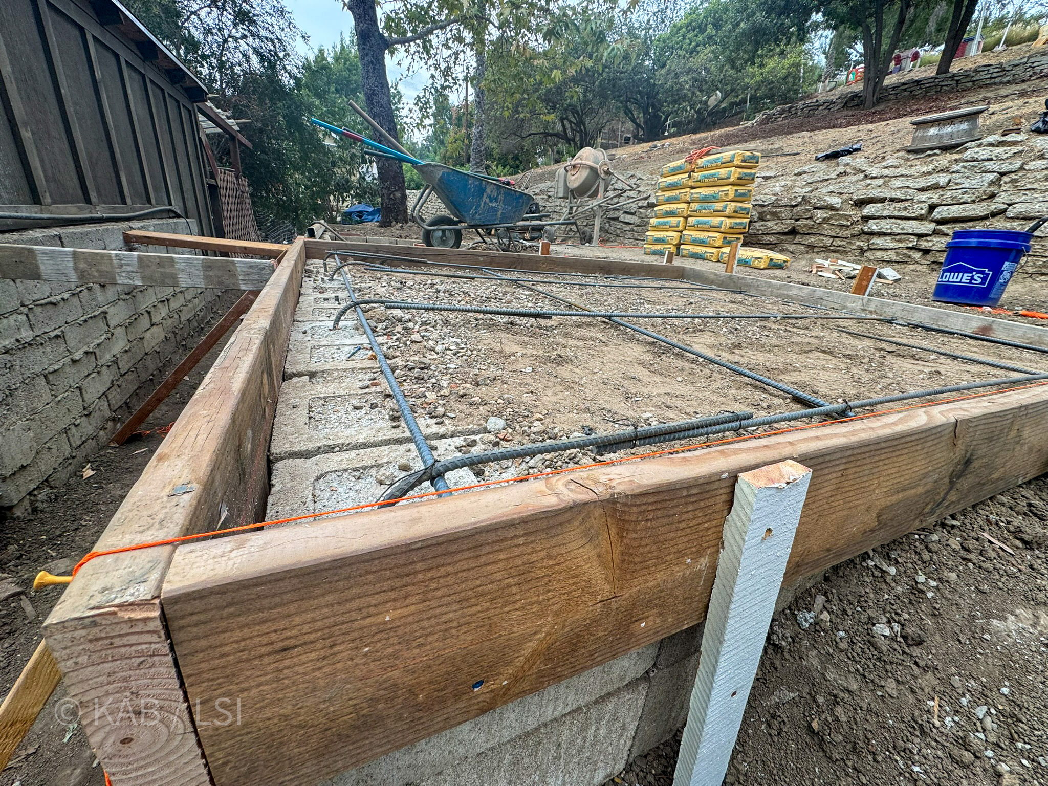 Framed concrete forms with rebar grid laid out on a hillside slope, with wheelbarrow, mixer, Quikrete stacks, and Lowe&rsquo;s bucket in background