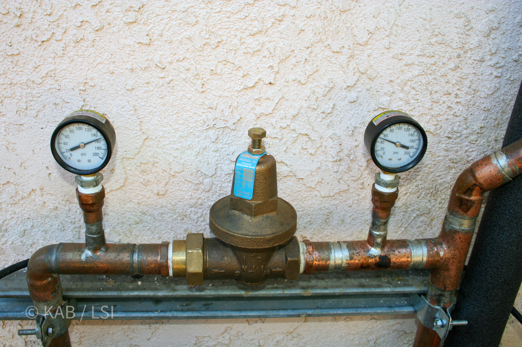 Dual pressure gauges on copper pipe flanking a brass Wilkins Model 70 pressure-regulating valve, showing 120 PSI incoming La Habra Heights city water regulated down to working pressure for the house