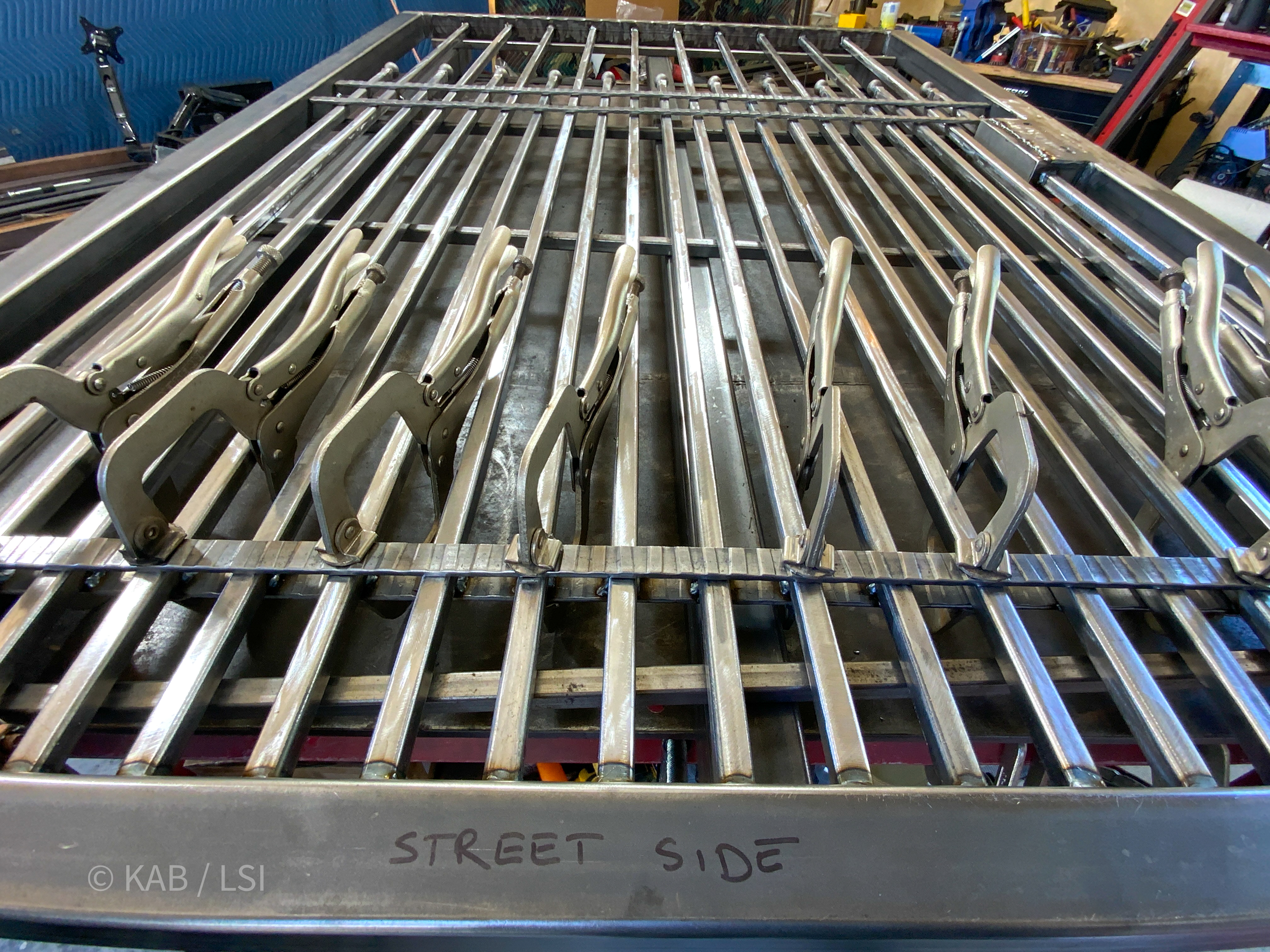 A coyote-proof wrought-iron fence panel under construction in a welding shop — lower pickets held to two inches on center, clamped for welding.
