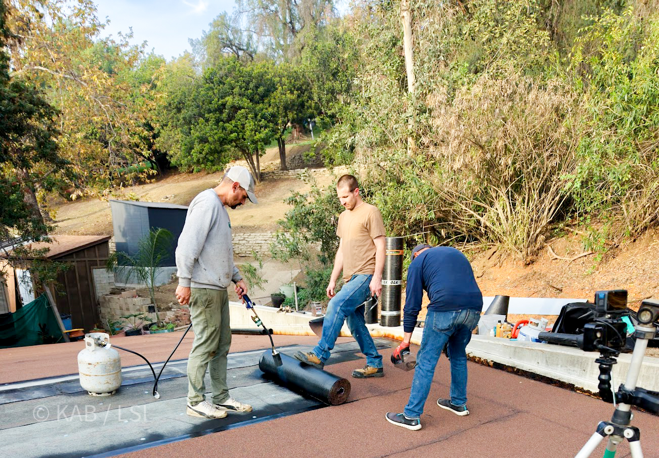 A La Habra Heights homeowner and his crew finish a torch-down roof on a hillside outbuilding before the storm arrives, viewed from above.
