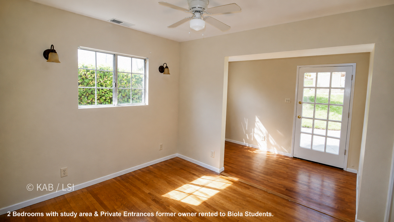 A bedroom with hardwood floors, beige walls, a ceiling fan, and two sconces flanking a window onto greenery. An opening at right leads to a light-filled study area with a glass-paned private-entrance door and a window looking onto a lawn.